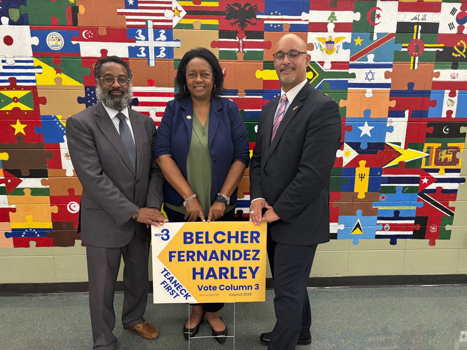 The Teaneck First slate with a yard sign in front of the world flags mural